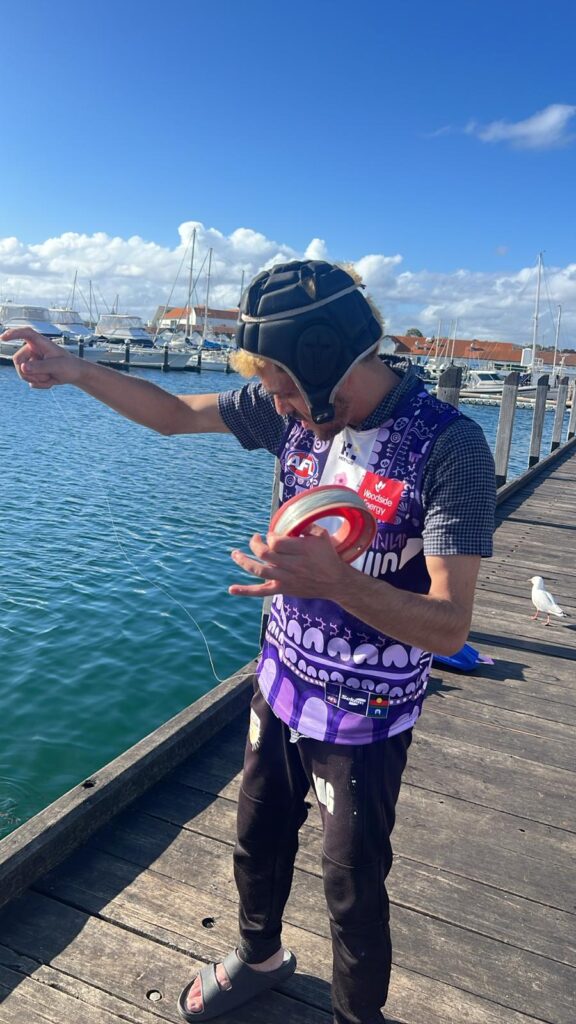 Hale Foundation participant in Fremantle Dockers AFL guernsey on a wooden jetty at the marina, handling fishing line with boats in the background